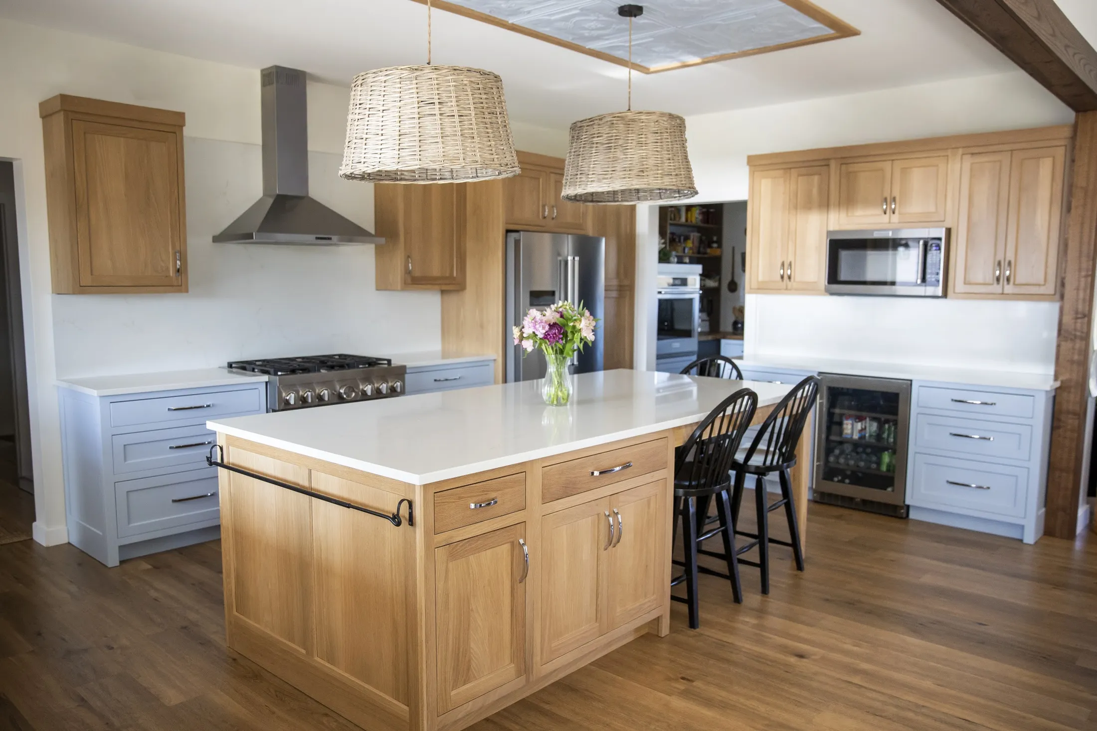 Oak island with black spindle stools and woven pendants under a coffered ceiling